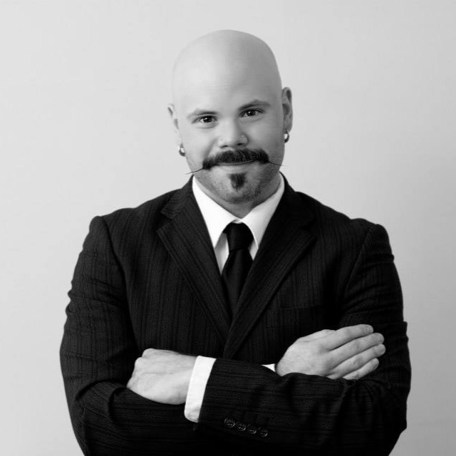 A man with a mustache and earrings poses confidently with his arms crossed, wearing a black pinstripe suit and a white shirt.