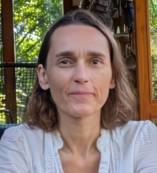 A close-up portrait of a woman with shoulder-length hair, wearing a white blouse, smiling softly while seated in a wooden setting with greenery in the background.